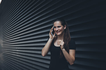 Porträt einer lächelnden jungen Frau, die mit einem Handy vor einer schwarzen Fassade telefoniert - CHAF001226