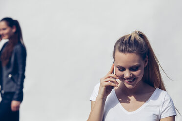 Porträt einer lächelnden jungen Frau, die mit einem Smartphone vor einem weißen Hintergrund telefoniert - CHAF001255