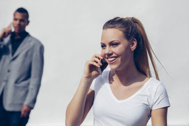 Porträt einer lächelnden jungen Frau, die mit einem Smartphone vor einem weißen Hintergrund telefoniert - CHAF001256