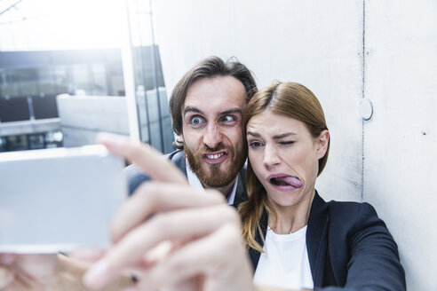 Porträt von zwei Geschäftsleuten, die Grimassen schneiden, während sie ein Selfie mit einem Smartphone machen - FMKF001742