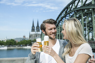 Germany, Cologne, happy young couple toasting with Koelsch glasses in front of Rhine River - FMKF001770