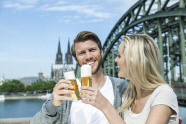 Germany, Cologne, happy young couple toasting with Koelsch glasses in front of Rhine River - FMKF001771