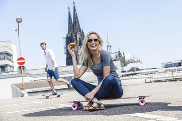 Deutschland, Köln, Porträt einer jungen Frau mit Bagel auf einem Skateboard sitzend - FMKF001779