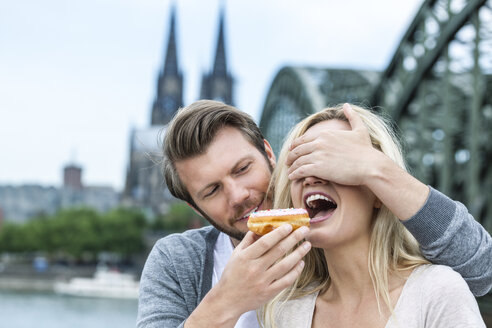 Germany, Cologne, young woman tasting a bagel while her boyfriend covering her eyes - FMKF001791