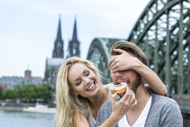 Germany, Cologne, young man tasting a bagel while his girlfriend covering his eyes - FMKF001792