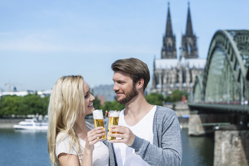 Deutschland, Köln, glückliches junges Paar stößt mit Kölschgläsern vor dem Rhein an - FMKF001799