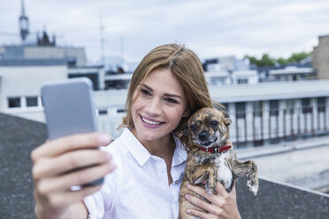 Porträt einer lächelnden jungen Frau, die ein Selfie mit ihrem Hund auf der Dachterrasse macht - FMKF001821