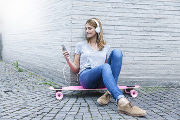 Deutschland, Köln, junge Frau auf rosa Skateboard sitzend und mit Kopfhörern Musik hörend - FMKF001803