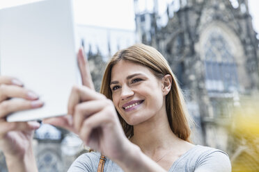 Germany, Cologne, portrait of smiling young woman taking a selfie in front of Cologne Cathedral - FMKF001810