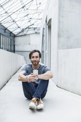 Portrait of smiling young man sitting on the floor with his smartphone - FMKF001830