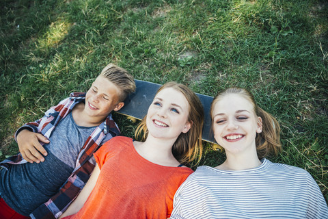 Drei glückliche Teenager-Freunde liegen auf einem Skateboard auf einer Wiese, lizenzfreies Stockfoto