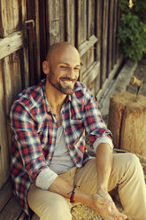 Porträt eines lächelnden Mannes mit kariertem Hemd, der vor einer Holzhütte sitzt - MAEF010921