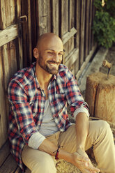 Portrait of smiling man wearing checked shirt sitting in front of wooden hut - MAEF010921