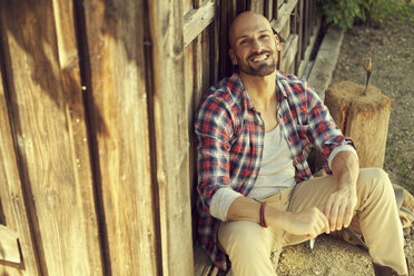 Portrait of man wearing checked shirt sitting in front of wooden hut - MAEF010922