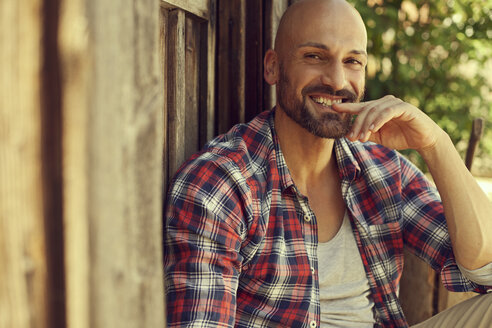 Porträt eines lächelnden Mannes mit kariertem Hemd, der vor einer Holzhütte sitzt - MAEF010924