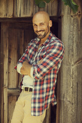 Porträt eines lächelnden Mannes mit kariertem Hemd, der vor einer Holzhütte steht - MAEF010925