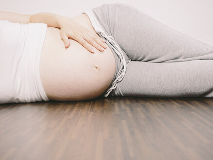 Schwangere Frau, die sich auf dem Holzboden ausruht und ihren Bauch hält - KRPF001621