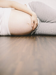 Schwangere Frau, die sich auf dem Holzboden ausruht und ihren Bauch hält - KRPF001620