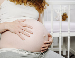 Pregnant woman in front of cot holding her belly - KRPF001615