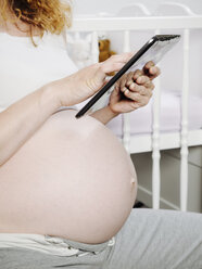 Pregnant woman in front of cot using a digital tablet - KRPF001605