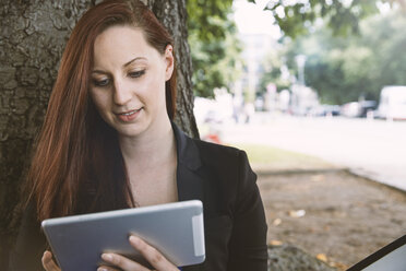 Junge Frau an einem Baum mit Blick auf ein digitales Tablet - STKF001389