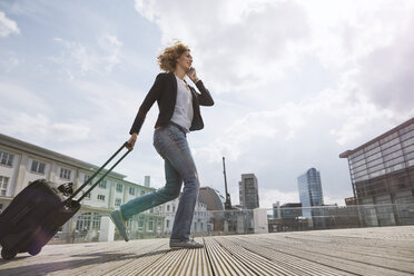 Woman on the phone walking with a rolling suitcase in the city - STKF001436