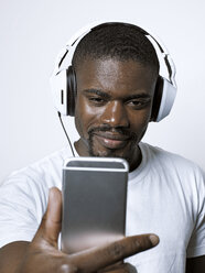Lächelnder junger Mann mit Kopfhörern, der auf sein Mobiltelefon schaut - STKF001440