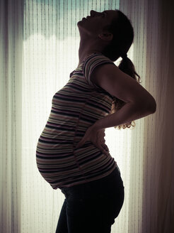 Schwangere Frau mit Rückenschmerzen vor hellem Fenster - KRPF001598