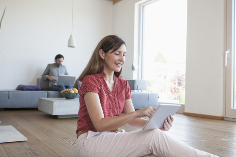 Ältere Frau benutzt digitales Tablet auf dem Boden im Wohnzimmer, Mann benutzt digitales Tablet im Hintergrund, lizenzfreies Stockfoto