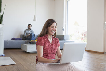 Reife Frau mit Laptop auf dem Boden im Wohnzimmer, Mann mit digitalem Tablet im Hintergrund - RBF003292