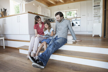 Happy family sitting on kitchen steps, parents reading book with daughter - RBF003361