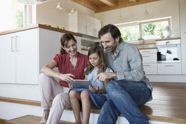 Happy family sitting on kitchen steps, daughter using digital tablet - RBF003309