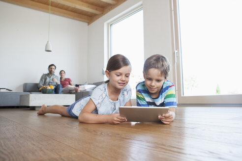 Bruder und Schwester liegen auf dem Boden und benutzen ein digitales Tablet, die Eltern schauen im Hintergrund zu - RBF003319