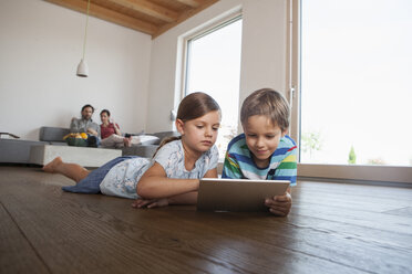 Bruder und Schwester liegen auf dem Boden und benutzen ein digitales Tablet, die Eltern schauen im Hintergrund zu - RBF003320