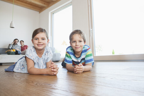 Bruder und Schwester liegen auf dem Boden, die Eltern schauen im Hintergrund zu - RBF003321