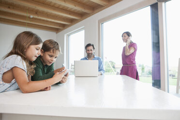 Bruder und Schwester benutzen ein digitales Tablet, die Eltern benutzen einen Laptop im Hintergrund - RBF003329