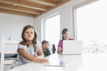 Little girl using digital tablet, mother and brother using laptop in background - RBF003330