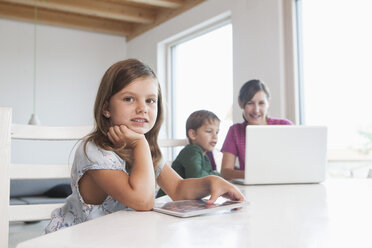 Kleines Mädchen mit digitalem Tablet, Mutter und Bruder mit Laptop im Hintergrund - RBF003331