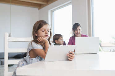Little girl using digital tablet, mother and brother using laptop in background - RBF003332