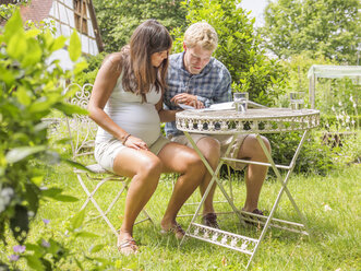 Young couple expecting baby, sitting in garden reading magazine - LAF001494