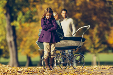 Kleines Mädchen mit verschränkten Armen vor einem Kinderwagen in einem Park mit Eltern im Hintergrund stehend - CHAF001079