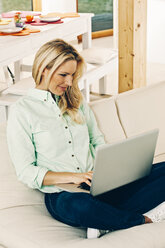 Smiling blond woman sitting on sofa using laptop - CHAF001261