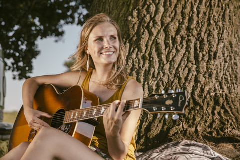 Junge Frau spielt Gitarre an einem Baum, lizenzfreies Stockfoto