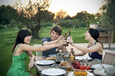 Happy friends sitting at garden table on a barbecue clinking beer bottles - TOYF001093