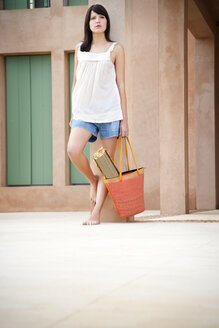 Young woman with bag and beach mat leaning against pillar - TOYF001145