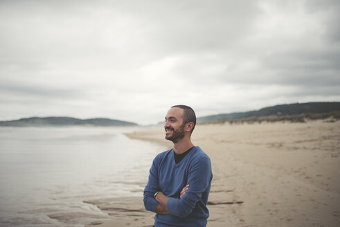 Spanien, Ferrol, Porträt eines lächelnden Mannes mit verschränkten Armen am Strand - RAEF000337