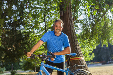 Porträt eines lächelnden Mannes, der mit seinem Fahrrad in einem Park steht - JUNF000427