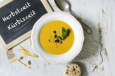 Selbstgemachte Kürbissuppe mit Kürbiskernen, Petersilie, einer Schiefertafel und einem Brötchen mit Oliven und Tomaten auf einem Holztisch - ODF001221