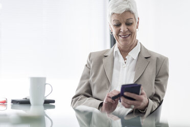 Lächelnde Geschäftsfrau im Büro, die auf ihr Mobiltelefon schaut - ZEF007403