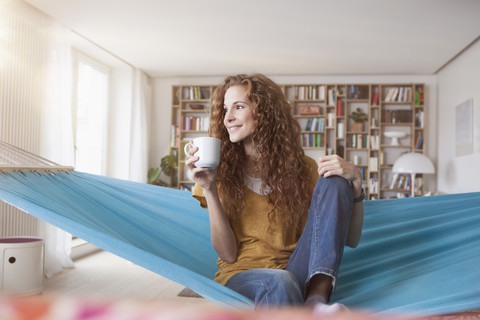 Lächelnde Frau zu Hause in der Hängematte sitzend mit einer Tasse Kaffee, lizenzfreies Stockfoto